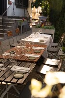 Un espacio de comedor al aire libre con mesas dispuestas para dos, con mesas de madera y sillas de metal.