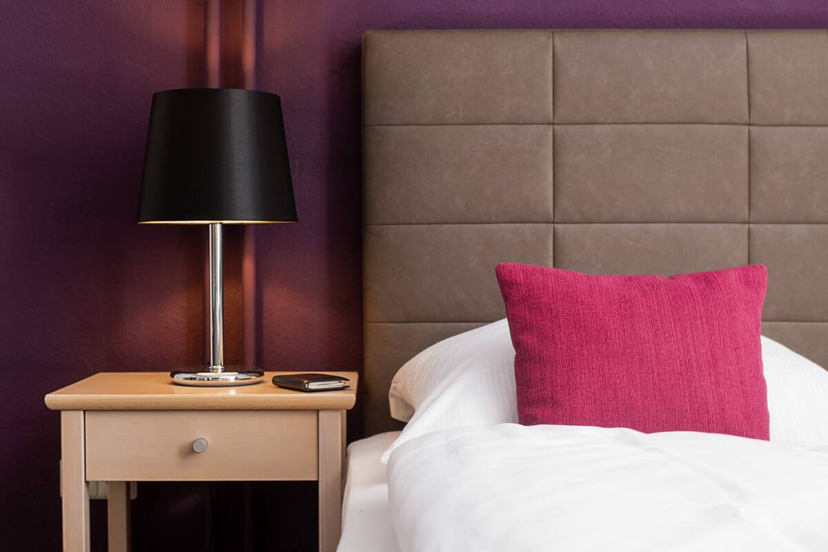 A neatly made bed with a white comforter and a single bright pink pillow, a black lamp on a wooden bedside table, and a brown upholstered headboard.