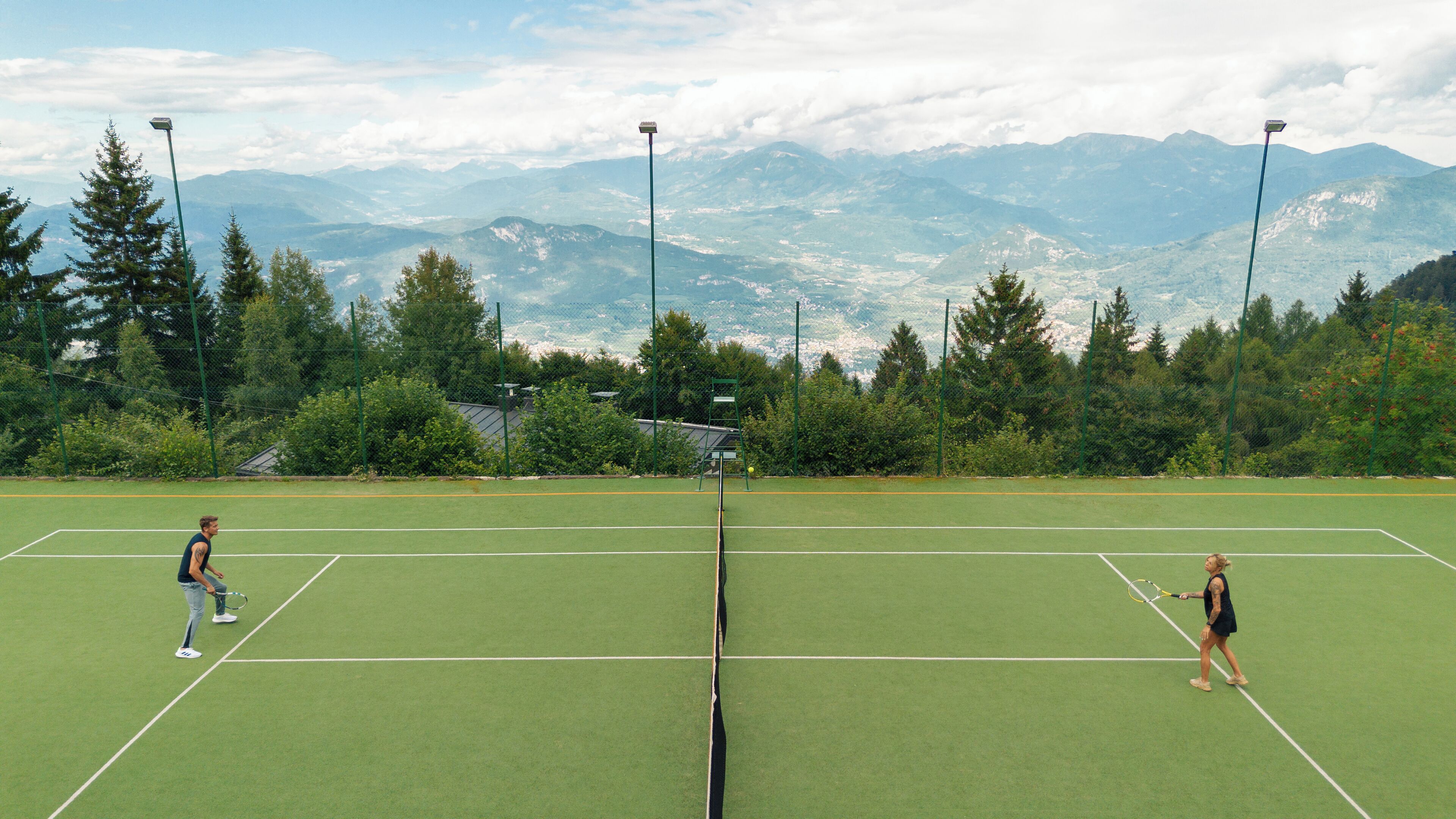 Cancha de tenis con dos jugadores, rodeada de montañas y árboles.