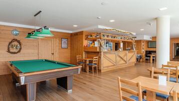 Une table de billard recouverte de feutre vert, des chaises en bois et un coin bar avec des étagères remplies de bouteilles et un téléviseur.