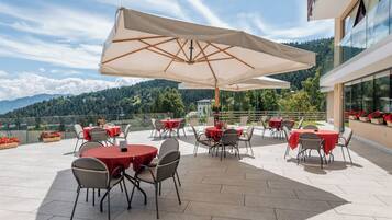 Una terrazza all'aperto con tavoli e sedie rotonde, ampi ombrelloni e una vista panoramica sulle montagne.