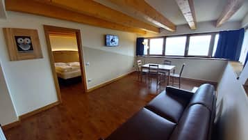 Um quarto aconchegante com piso de madeira, uma cama, uma TV, uma mesa de jantar com cadeiras e um sofá.
