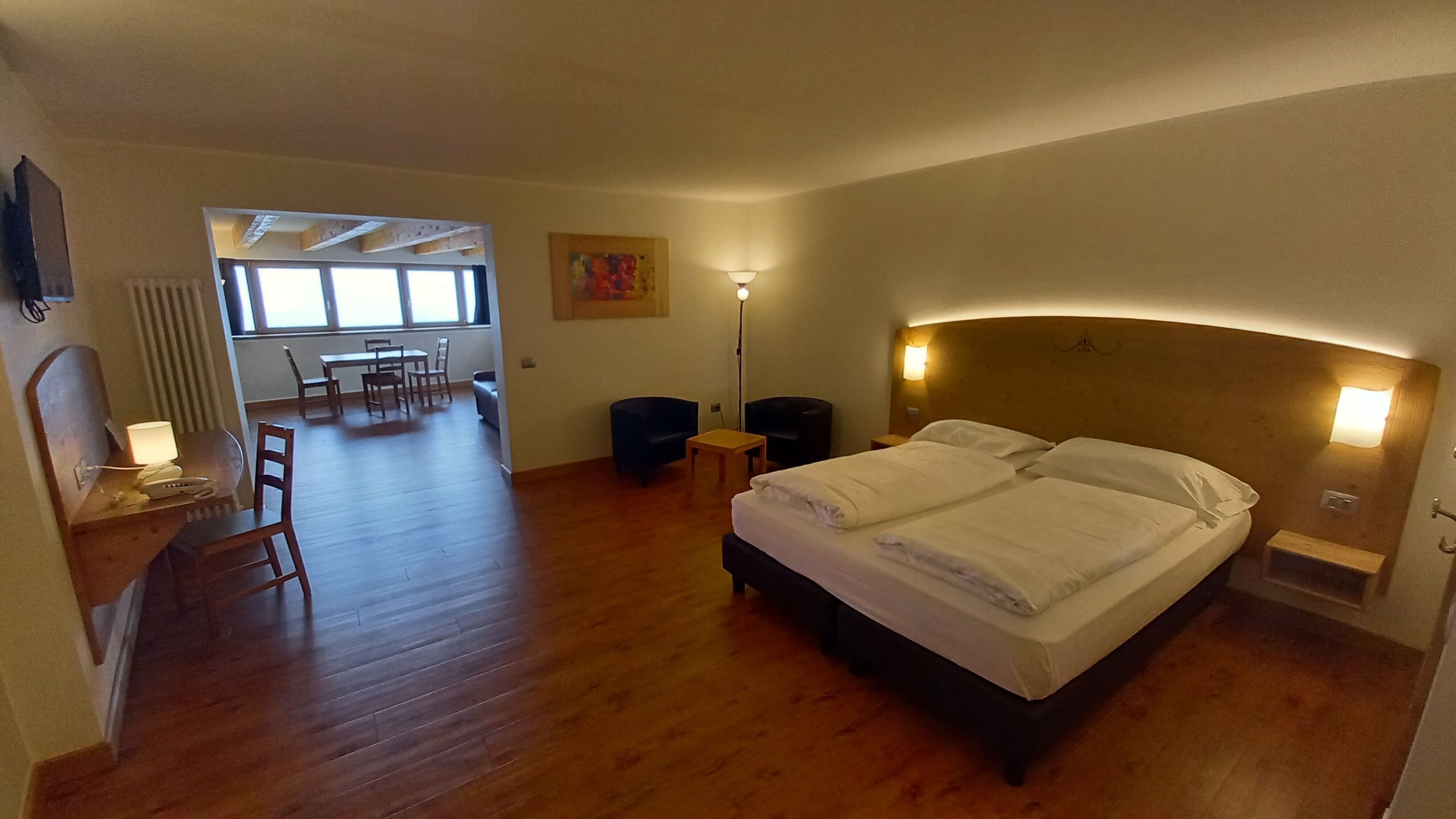 A hotel room with a large bed, a desk with a lamp, a chair, and a dining area with a table and chairs.