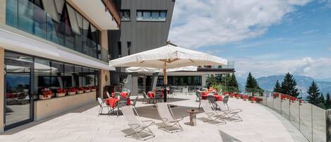 En moderne udendørs terrasse med liggestole og parasoller, med udsigt over et bjergrigt landskab.