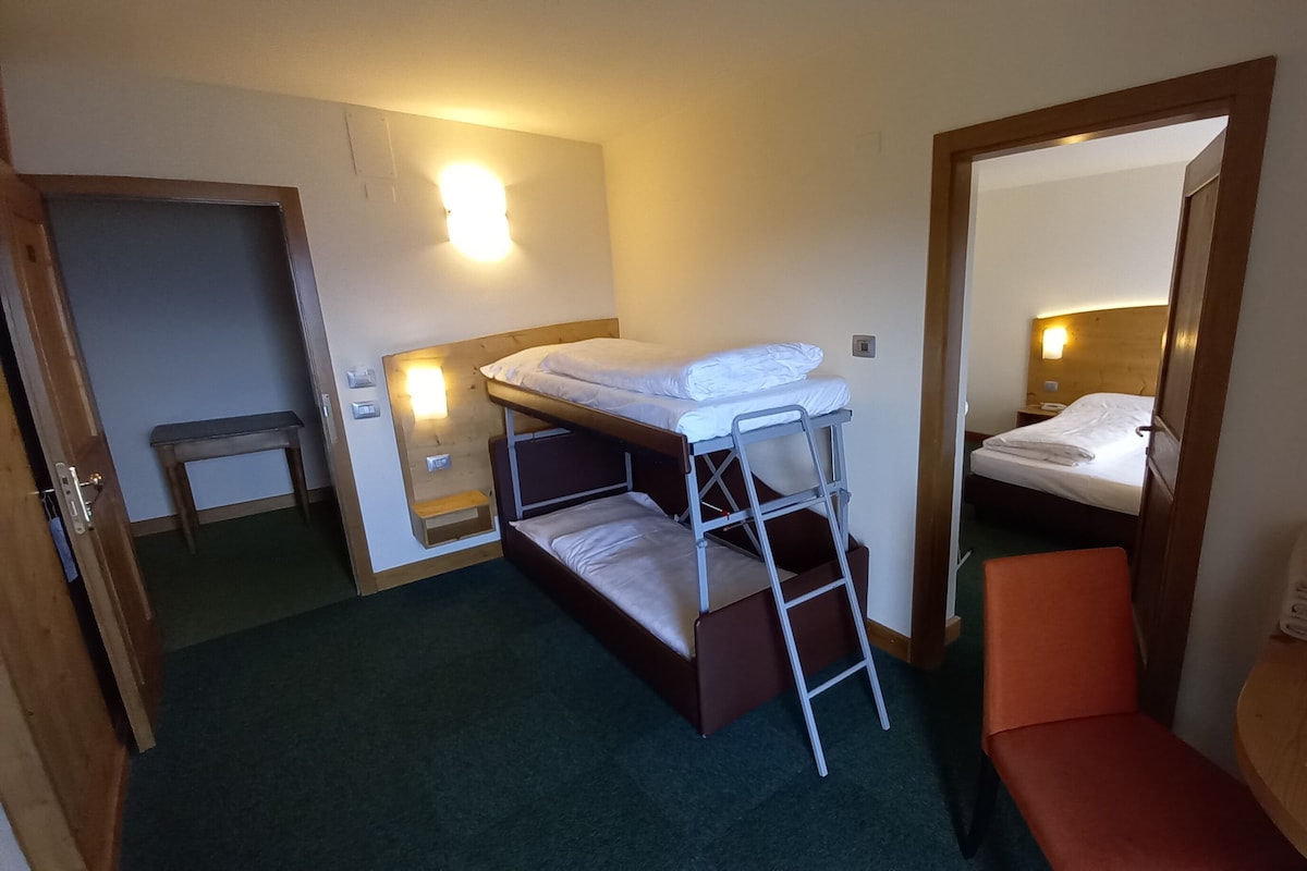 A compact hotel room with a bunk bed, a small bench, a bedside table, and a mirror.