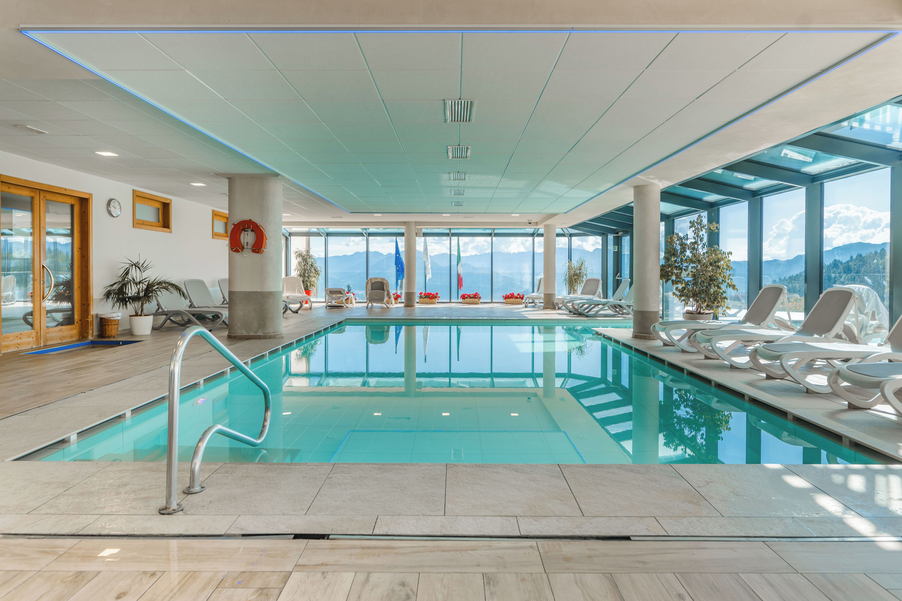 A large indoor swimming pool with clear glass walls, lounge chairs, and a view of mountains.