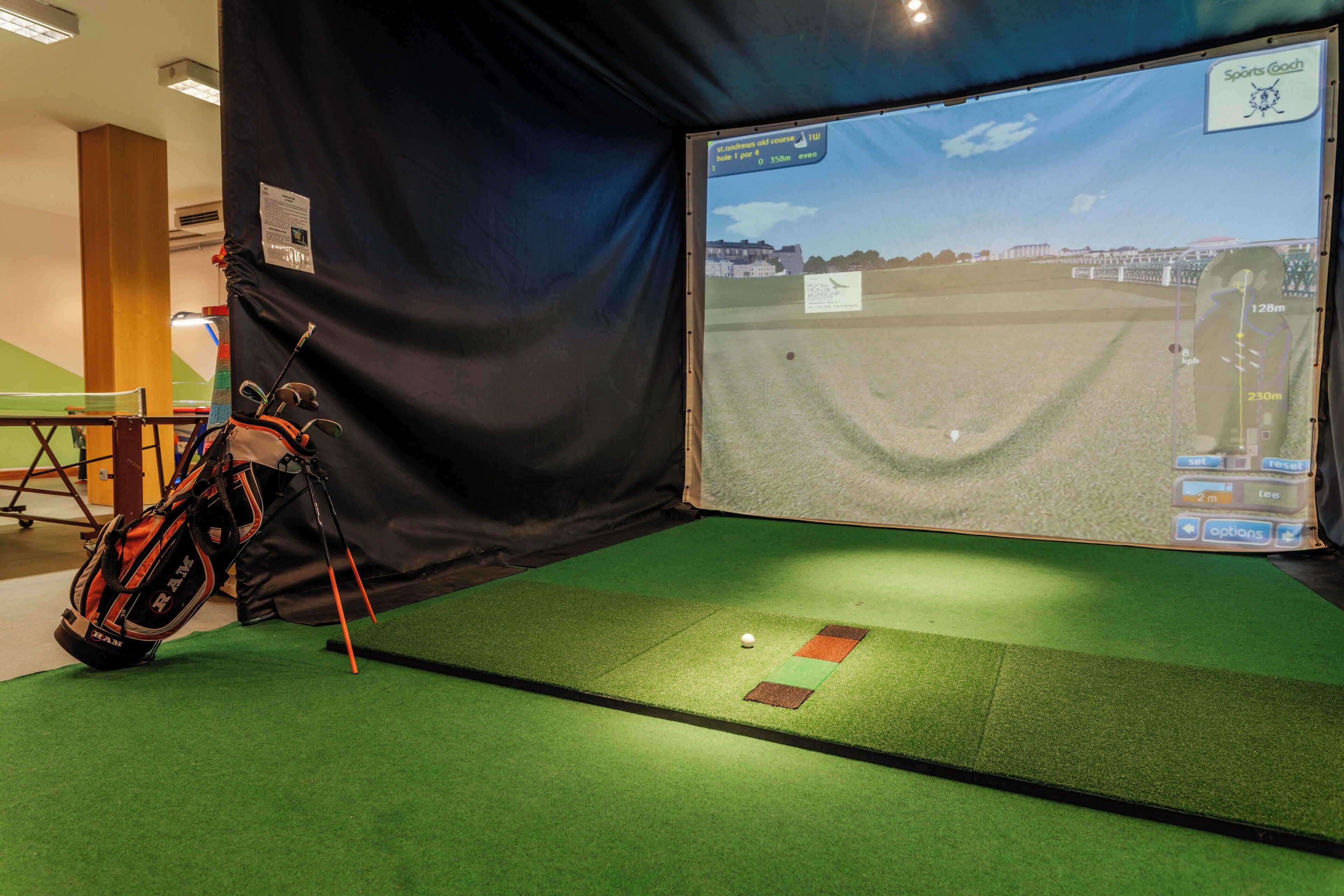 Ein Golf-Simulationssystem mit Bildschirm, der einen Golfplatz zeigt, einer Golfbag und einem Putting-Green.