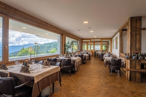 A restaurant with multiple tables, wooden flooring, and large windows offering a mountain view. - Hotel Monte Bondone (Trento)