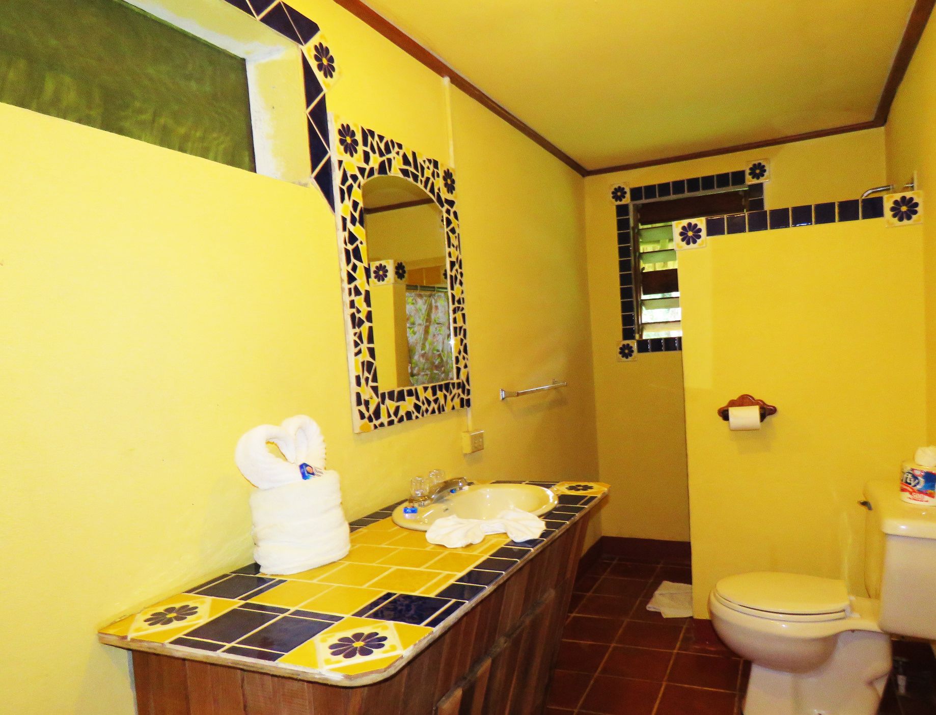 Un baño con pared amarilla, lavamanos con azulejos, un espejo y un inodoro.