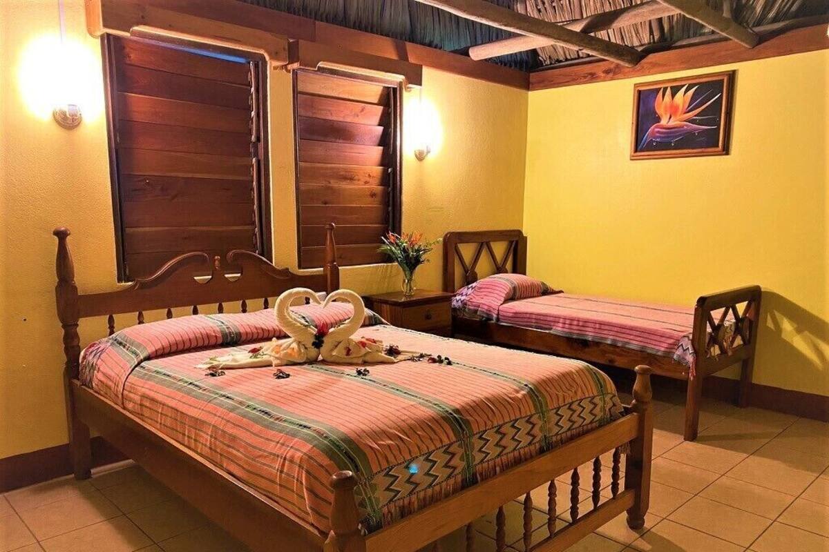 A room with a large bed, a smaller bed, and a chair. There are two windows with wooden shutters, a wall-mounted light, and a painting.