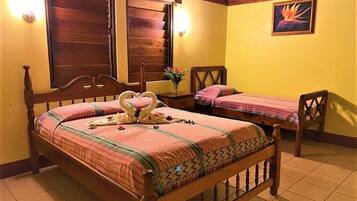 A room with a large bed, a smaller bed, and a chair. There are two windows with wooden shutters, a wall-mounted light, and a painting.