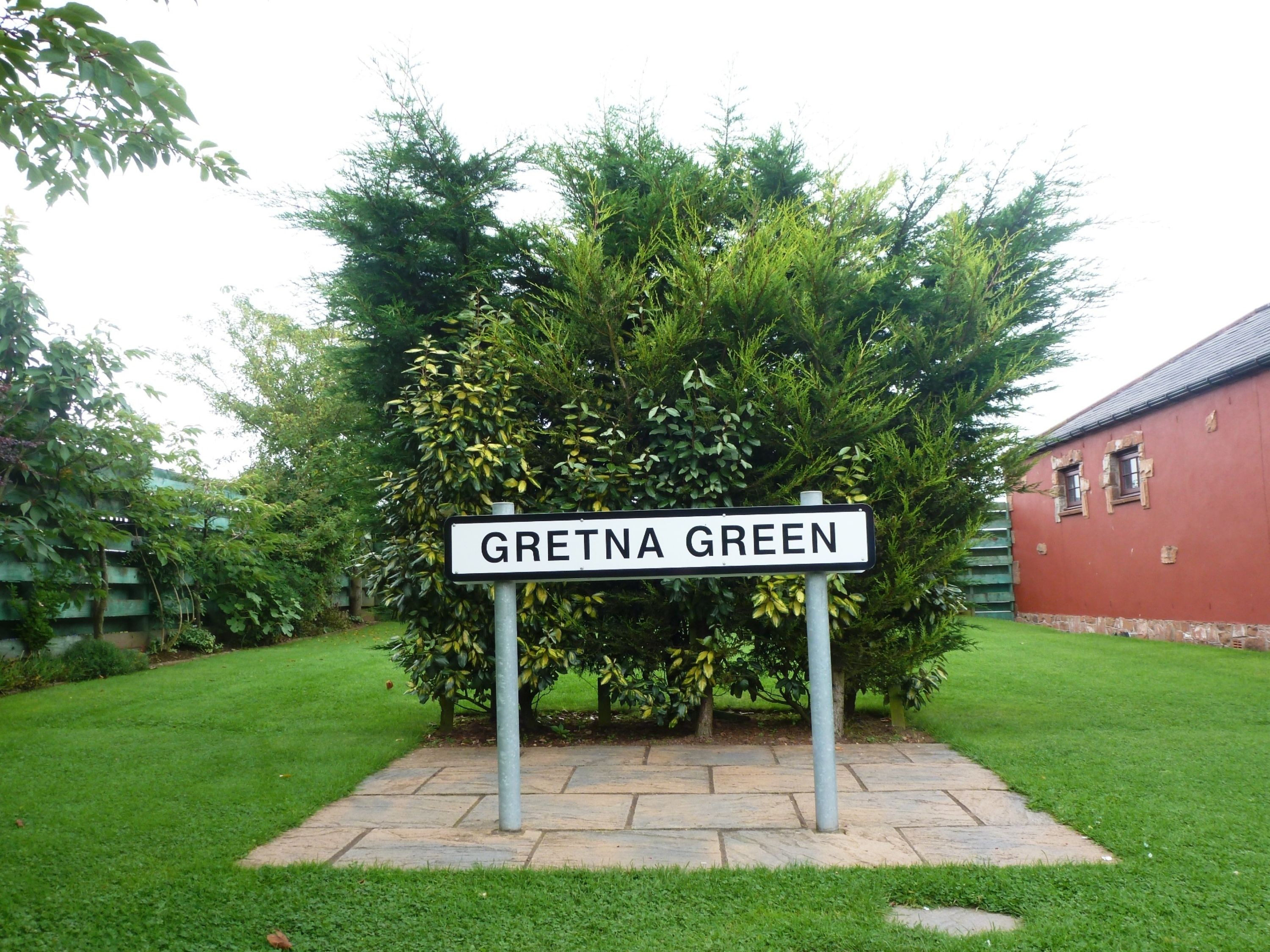 Ein Straßenschild mit der Aufschrift 'Gretna Green' in einem Garten, im Hintergrund ein rotes Gebäude.