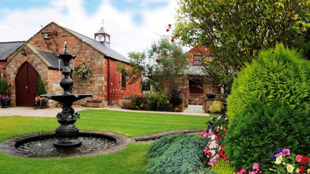 A stone building with a red door and a bell tower, surrounded by a well-maintained garden with a fountain and colorful flowers. - The Mill Forge (Lockerbie)