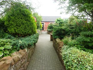 A paved pathway flanked by stone walls and lush greenery, leading to a red building with a dark roof.