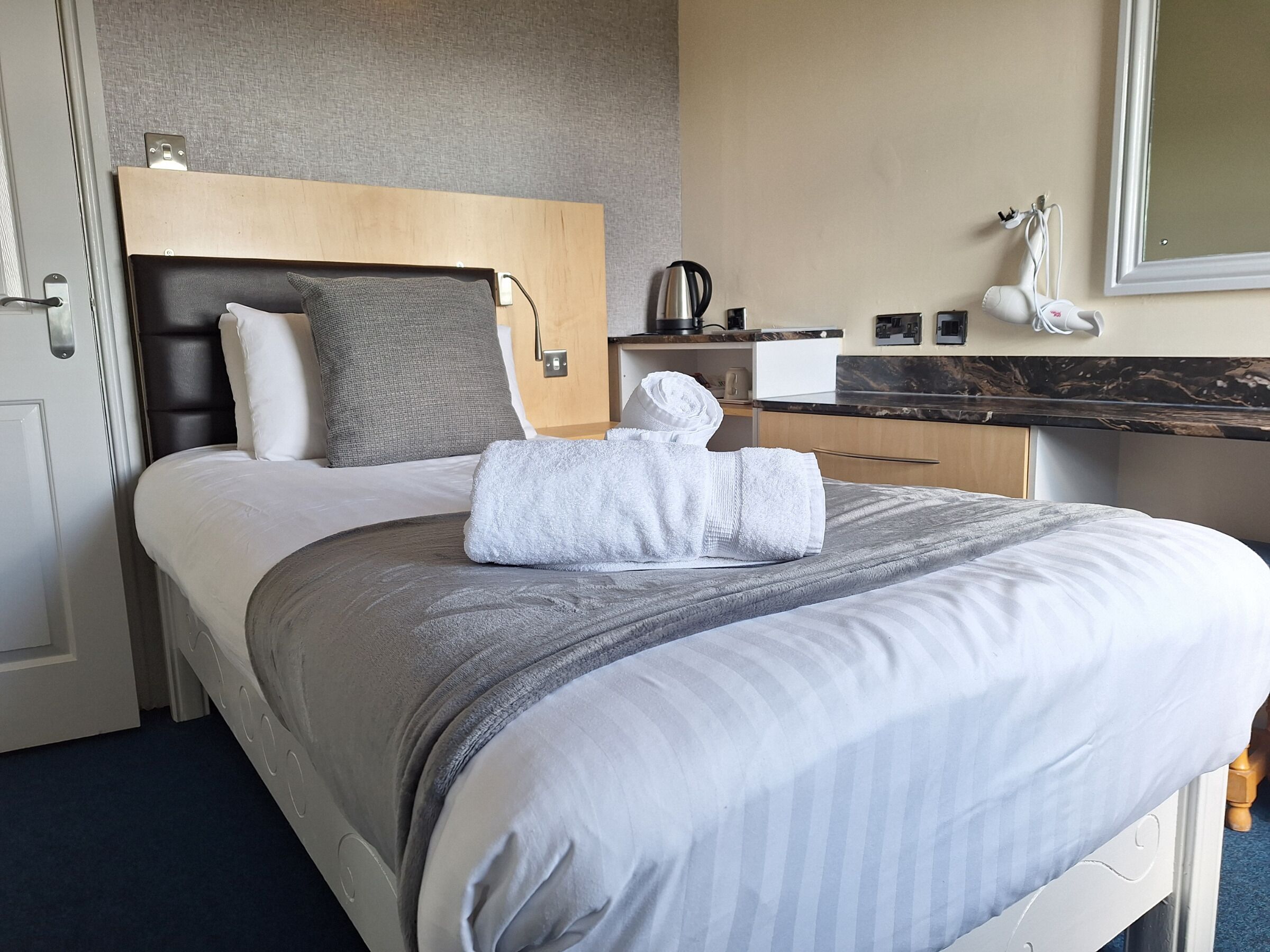 A hotel room with a bed, a wooden headboard, a bedside table with a kettle, a mirror, and a small desk with a lamp.
