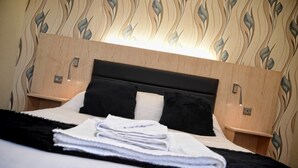 A neatly made bed with a black and white duvet, a folded white towel, and a black throw blanket. The room features a wooden headboard with a light fixture above and two bedside tables with lamps. - The Mill Forge (Lockerbie)