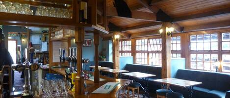A bar area with wooden beams, shelves with bottles, glasses, and stools. There are tables and a sofa in the seating area.