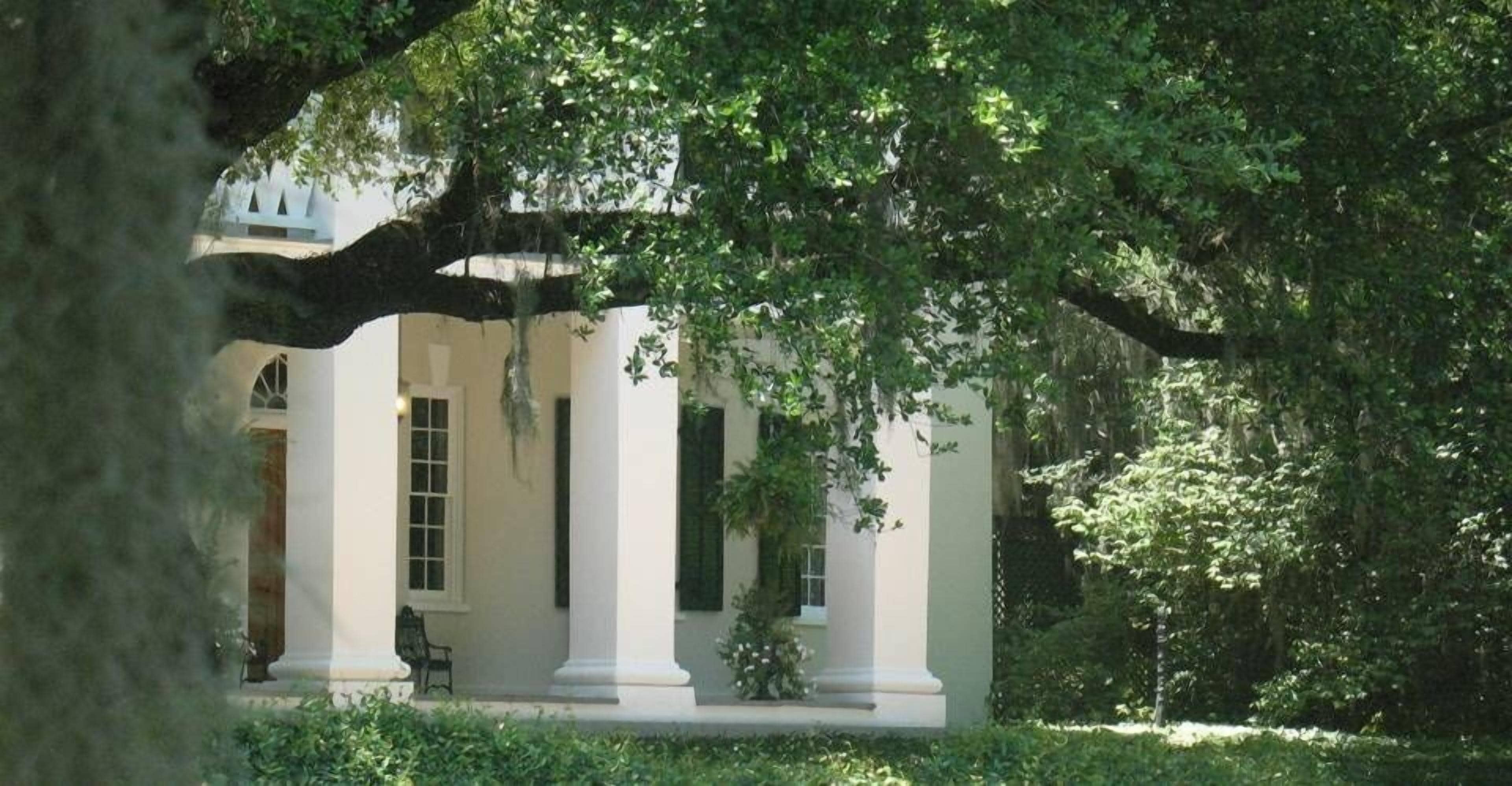 A building with white columns and a covered porch, surrounded by lush greenery.