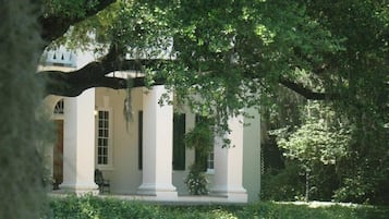A building with white columns and a covered porch, surrounded by lush greenery.