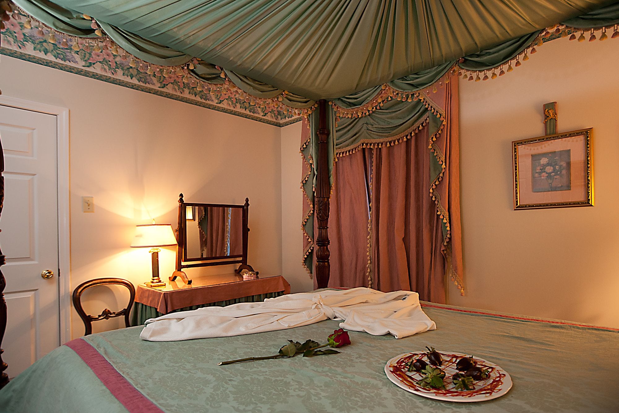 A bedroom with a bed, a bedside table, a mirror, a framed picture on the wall, and a rose on the bed.