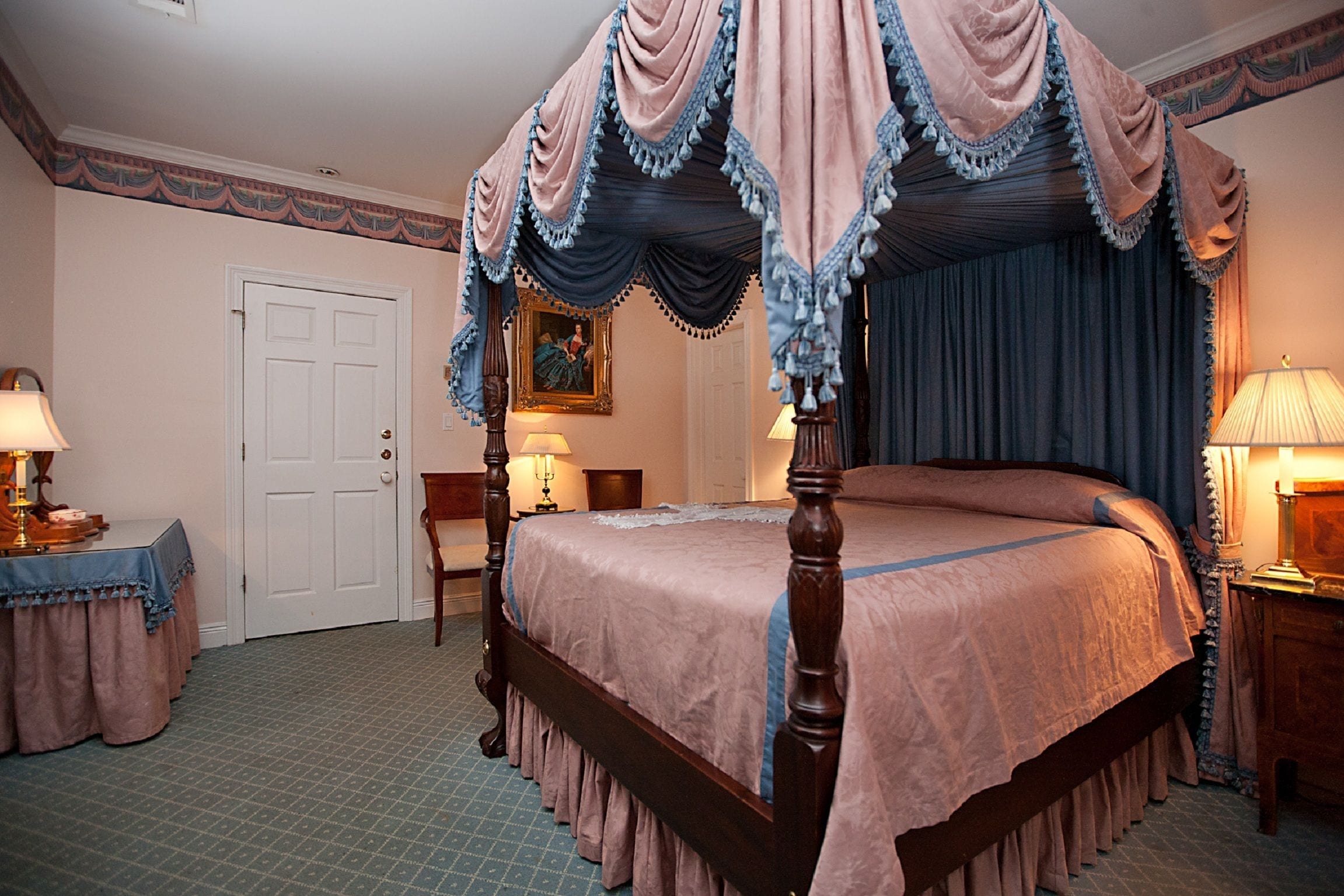 A four-poster canopy bed with a wooden frame and a pink bedspread. There is a wooden nightstand with a lamp on it, and a chair with a table beside the bed. A door is visible in the background.