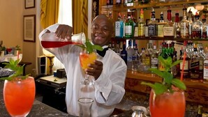 Una zona de bar con un bartender sirviendo una bebida en un vaso, una estantería con varias botellas de licores y dos vasos con una bebida roja sobre el mostrador.
