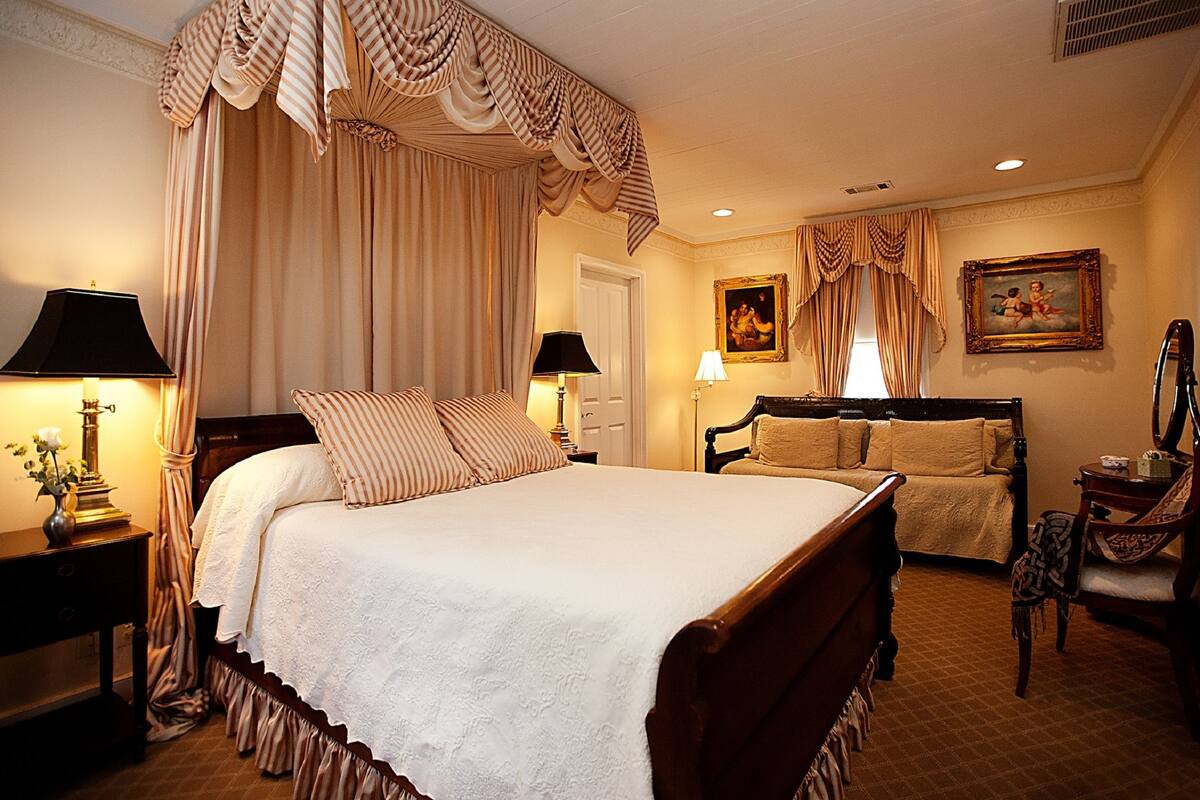 A traditional bedroom with a four-poster bed, a sitting area with a sofa, and a mirror.