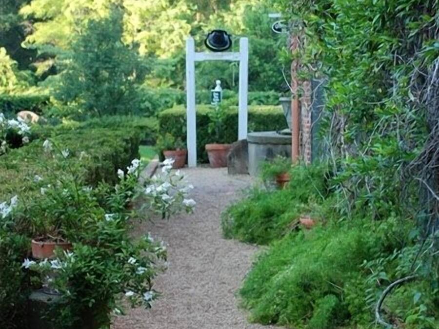 A garden pathway leading to a white archway with a sign, surrounded by lush greenery and potted plants.