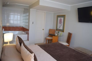 A hotel room with a bed, a desk, a chair, a television, a bathroom with a shower, and a framed picture on the wall.