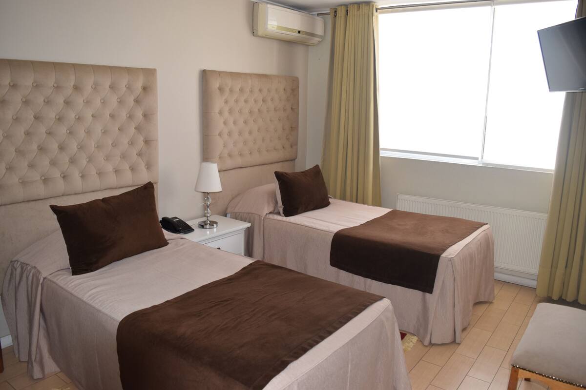 A hotel room with two beds, a bedside table with a lamp, a TV, and a window with curtains.