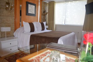 A hotel room with a bed, a nightstand, a lamp, a window with curtains, and a vase with a red flower.