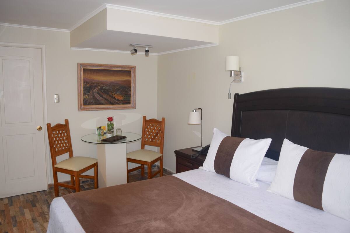 A hotel room with a bed, two chairs, a small table, a lamp, and a painting on the wall.