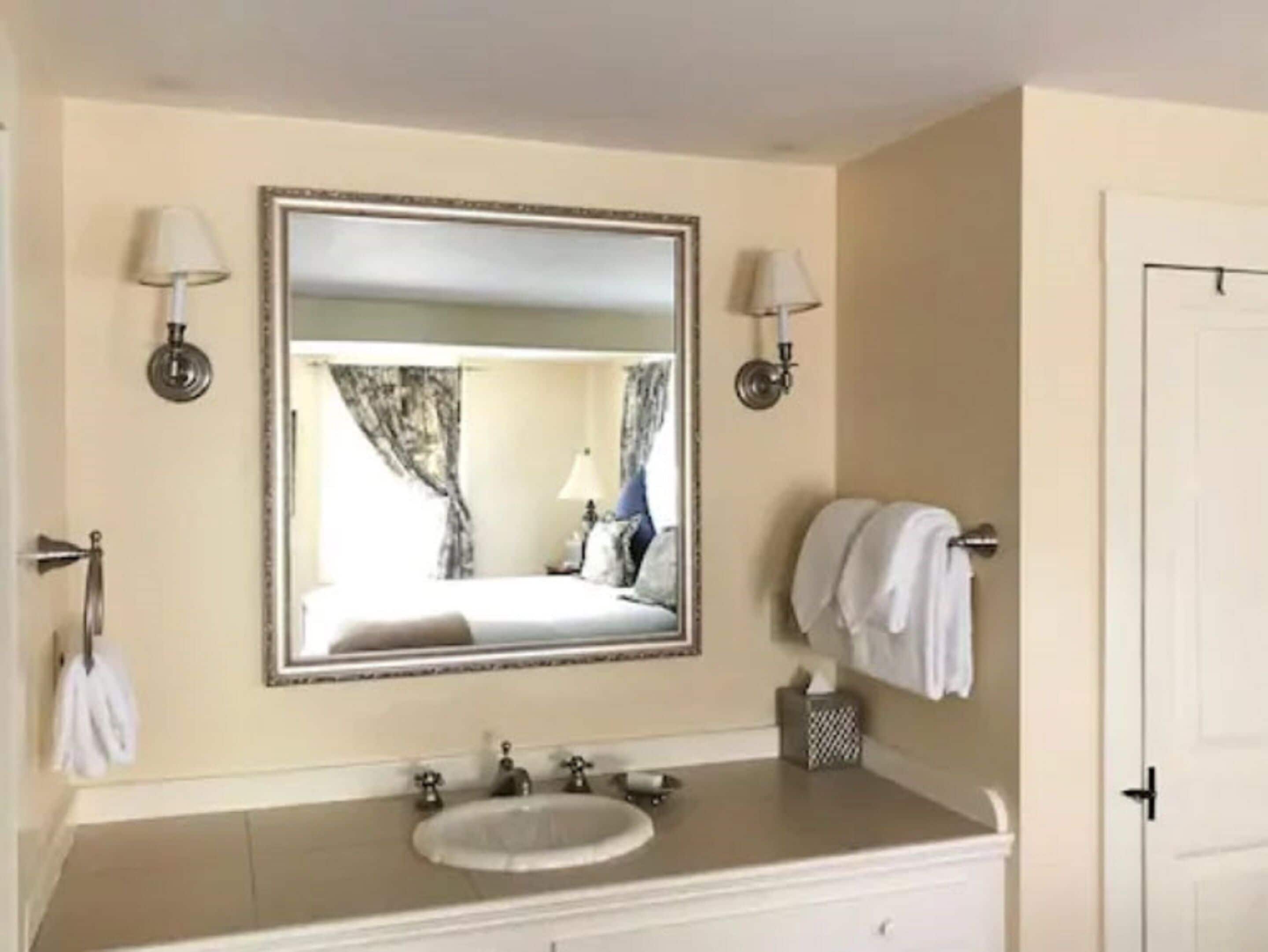 A bathroom with a large mirror, two wall-mounted lamps, a sink, and a towel rack.