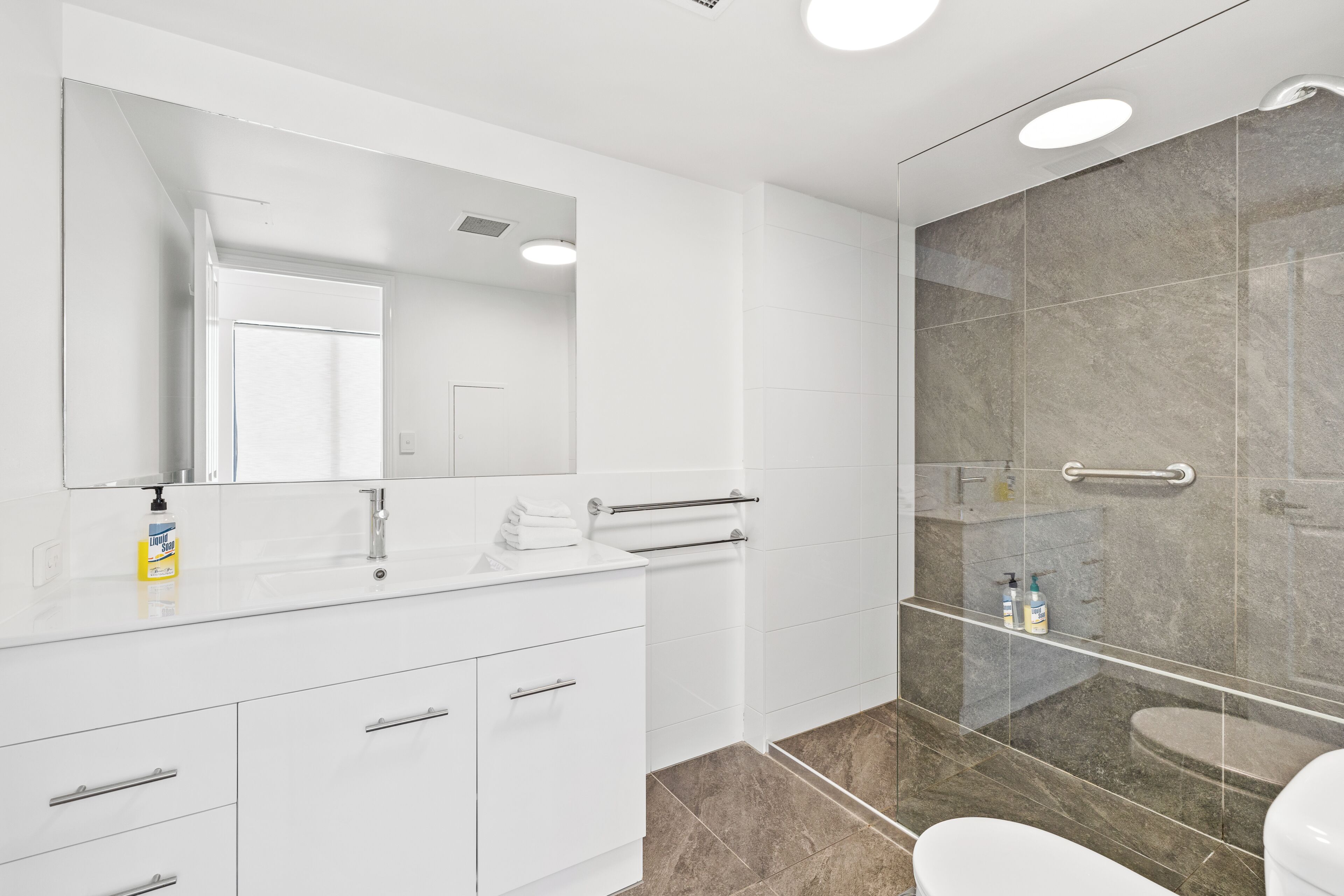 A modern bathroom with a white vanity, a large mirror, a glass shower enclosure, and a toilet.