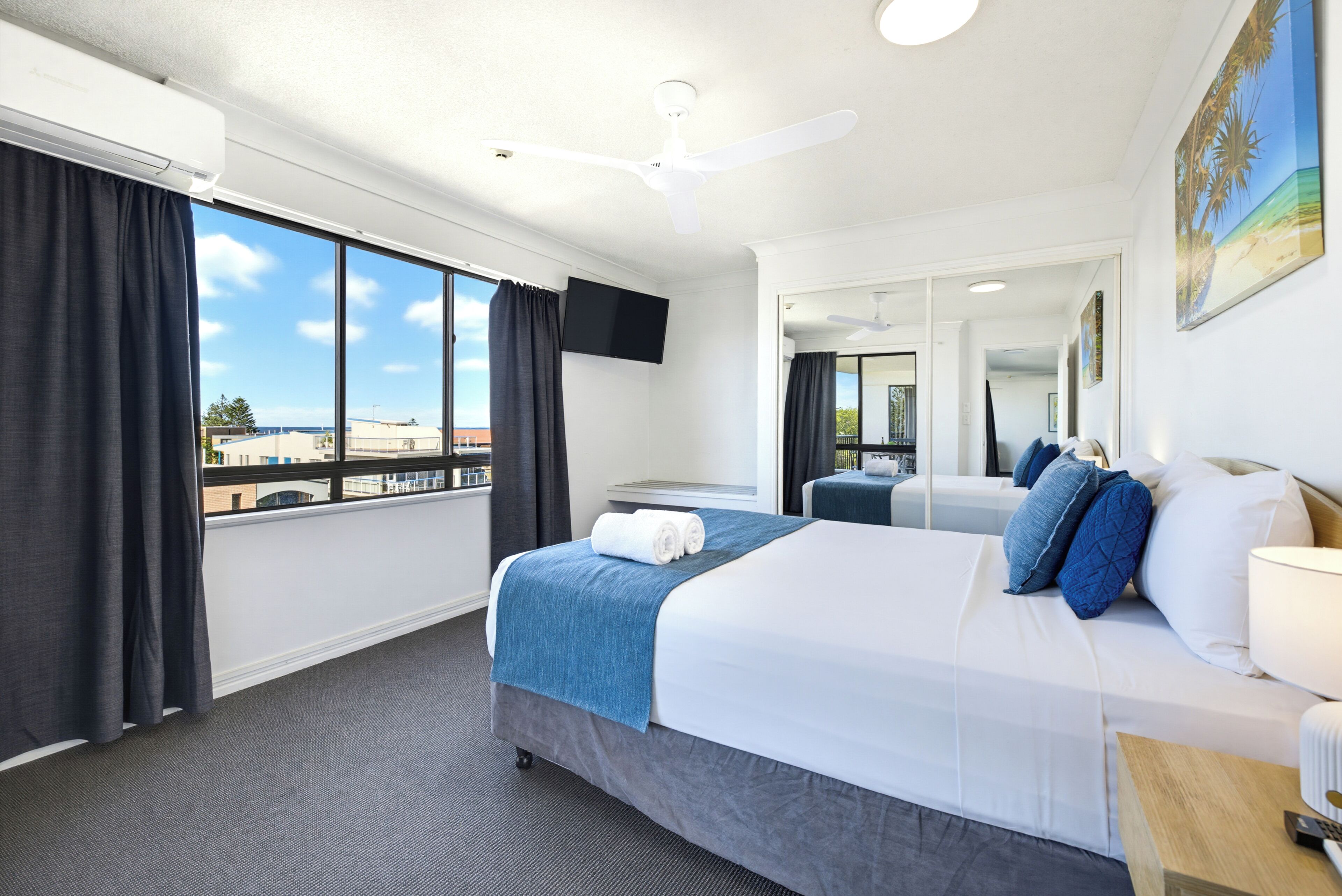 A hotel room with a large bed, a flat-screen TV, a ceiling fan, and a view of the cityscape.