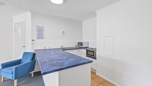 A small kitchenette with a microwave, oven, and a sink. There is a blue chair and a clock on the wall. - Belaire Place Motel Apartments (Caloundra)