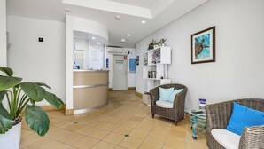 A reception area with a counter, seating, and a wall-mounted shelf with books and decorative items. - Belaire Place Motel Apartments (Caloundra)