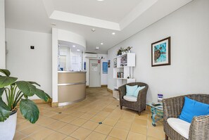 A reception area with a counter, seating, and a wall-mounted shelf with books and decorative items. - Belaire Place Motel Apartments (Caloundra)
