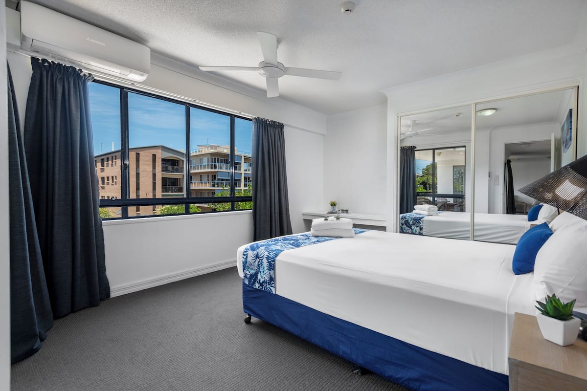 Une chambre d’hôtel avec un grand lit, un bureau, une chaise, un ventilateur de plafond et une fenêtre avec des rideaux.