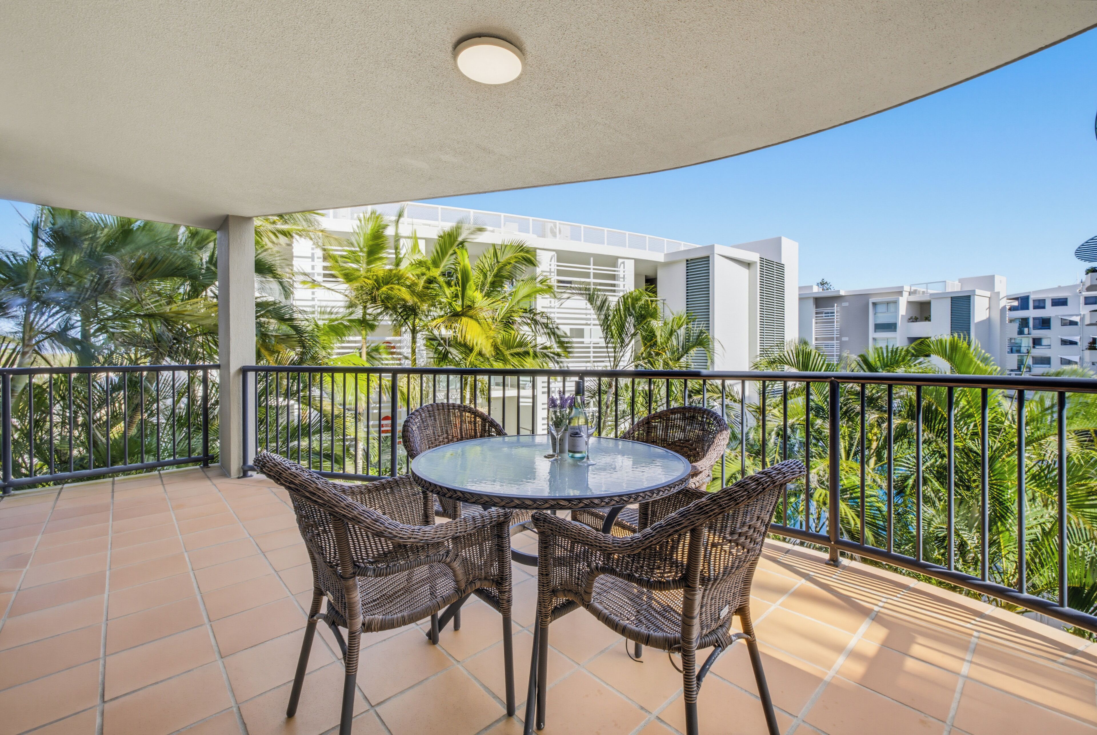 Een ronde tafel met rieten stoelen op een balkon, uitzicht op palmbomen en gebouwen.