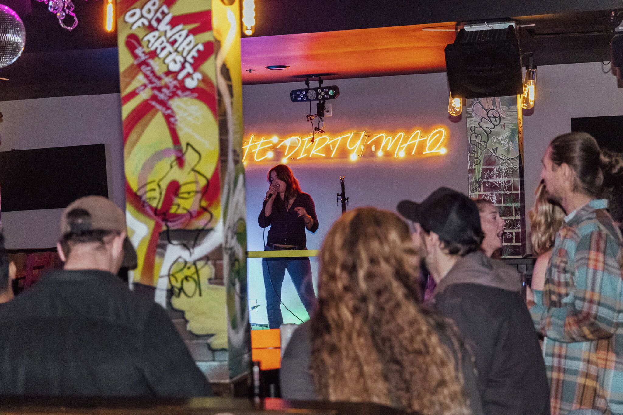 Una persona actuando en el escenario con un micrófono, un público frente a ella y un letrero de neón al fondo.