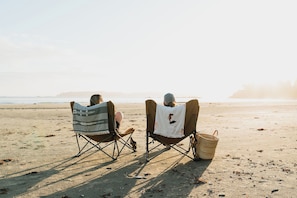 Two people seated in beach chairs facing the ocean, with a beach and distant landforms in the background. - The Maq Hotel (Tofino)