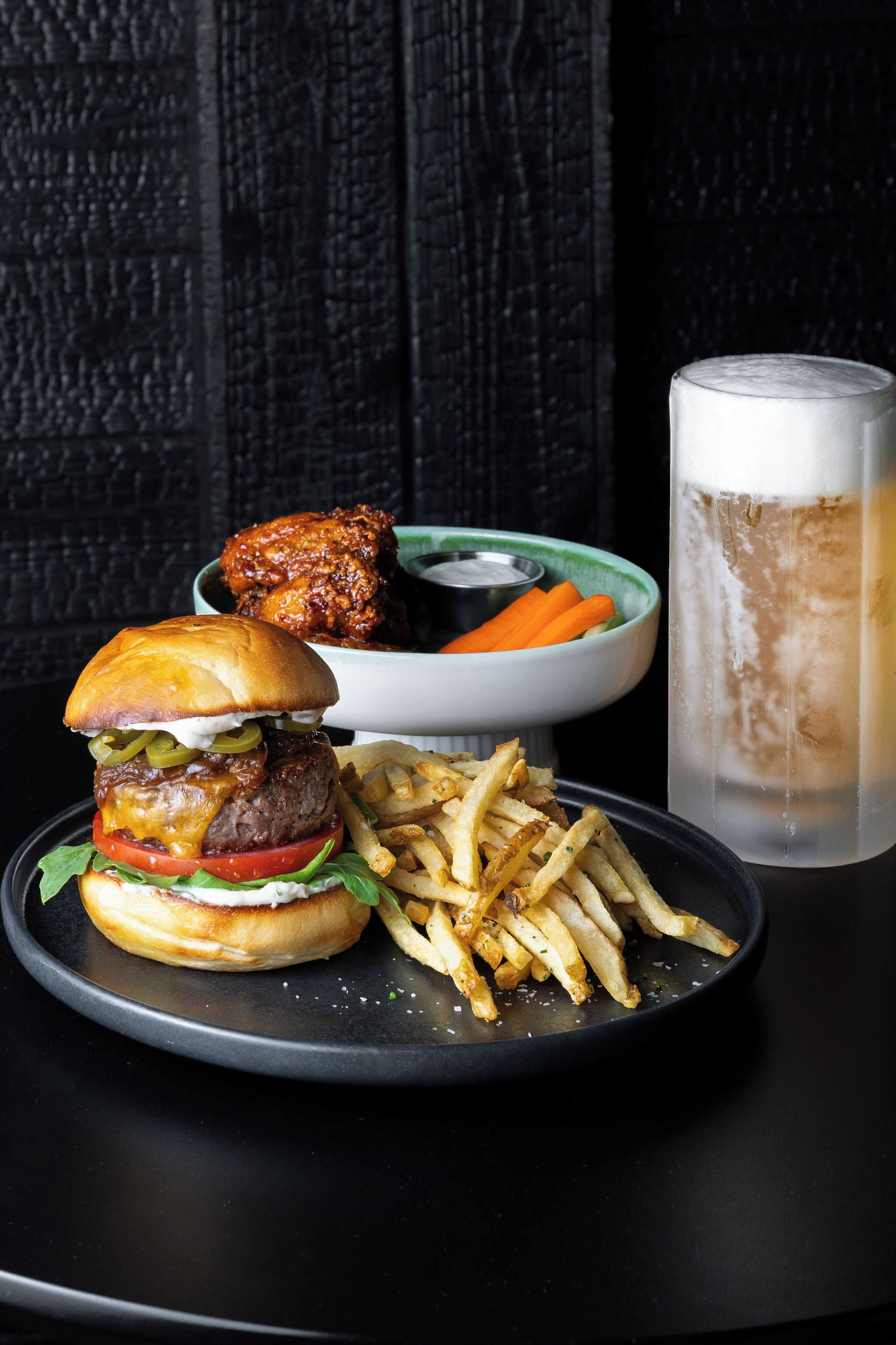 Een maaltijd bestaande uit een hamburger, frietjes en een glas bier op een donker bord.