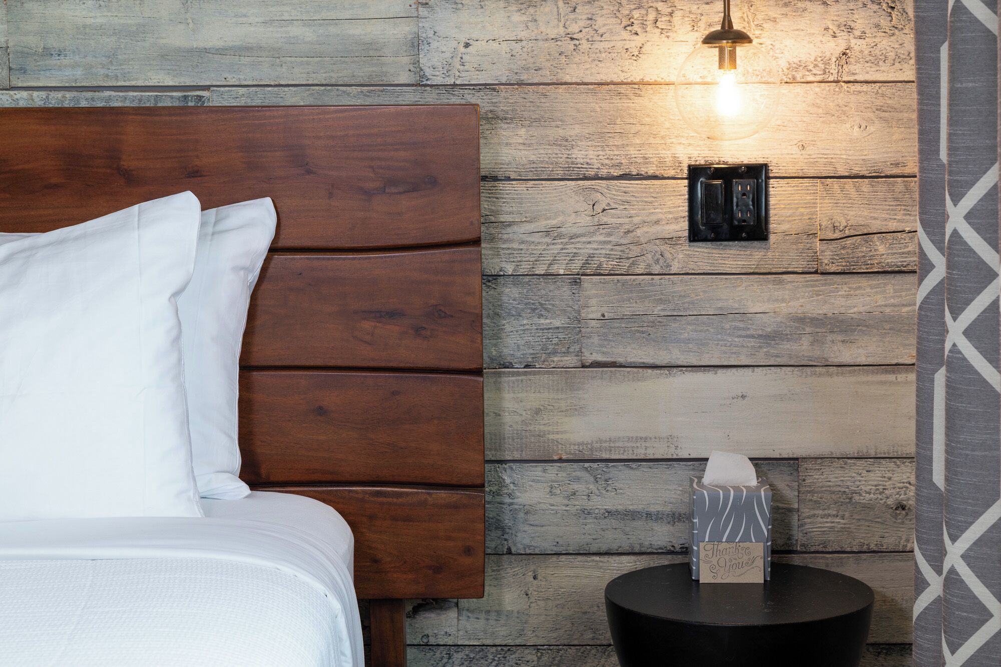 Un dormitorio con cabecera de madera, una mesita de noche con un dispensador de pañuelos y una lámpara fijada a la pared.