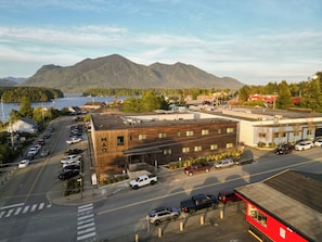A parking lot with multiple vehicles, a building with 'MAQ' on its facade, and a mountainous landscape in the background. - The Maq Hotel (Tofino)