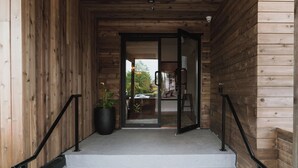 Fachada de madera con puerta de vidrio, una planta en maceta y un espacio exterior visible con un coche y vegetación.
