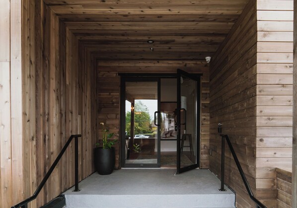 Fachada de madera con puerta de vidrio, una planta en maceta y un espacio exterior visible con un coche y vegetación.