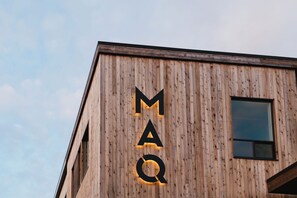 Un edificio con fachada de madera y una gran letra iluminada 'M' seguida por 'A' y 'Q' en la fachada.