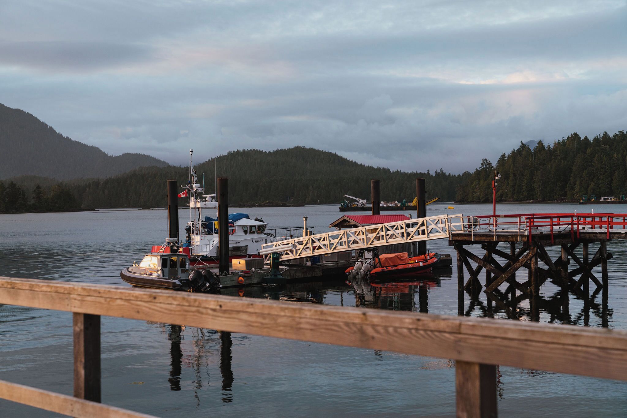 A dock with a boat moored alongside, a pier extending into calm water, and a forested hill in the background.