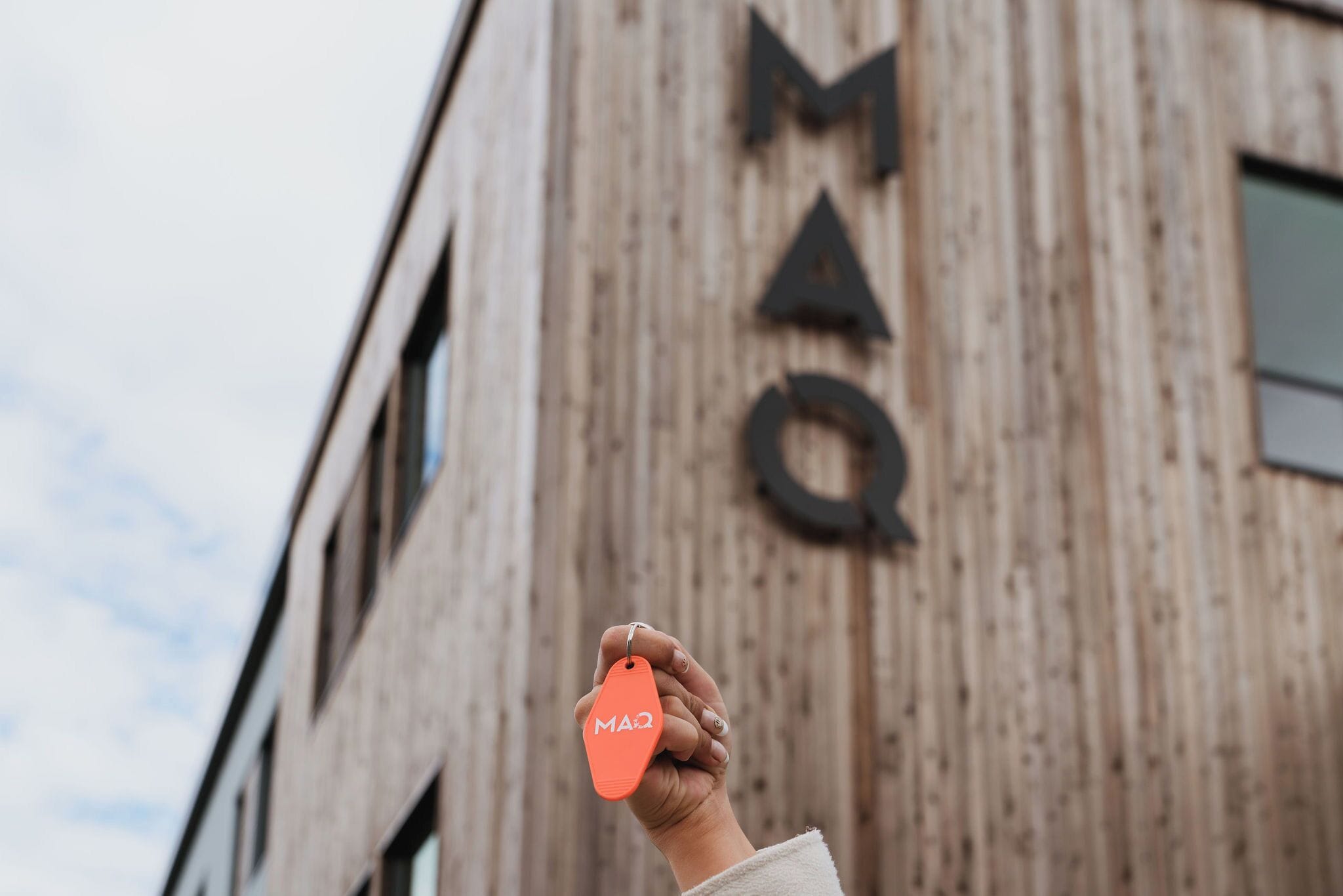 A hand holding a keychain with the letters 'MAQ' against a building with a wooden facade and large letters 'MQ' on it.